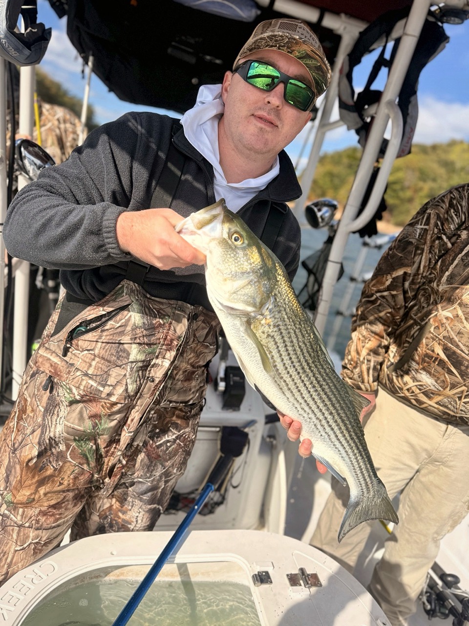 Lake Lanier Striper Fishing Report – October 24, 2026 | Tough Conditions, Quality Fish & Fall Migration Family fishing trip guest holding a Lake Lanier striped bass beside the bait tank
